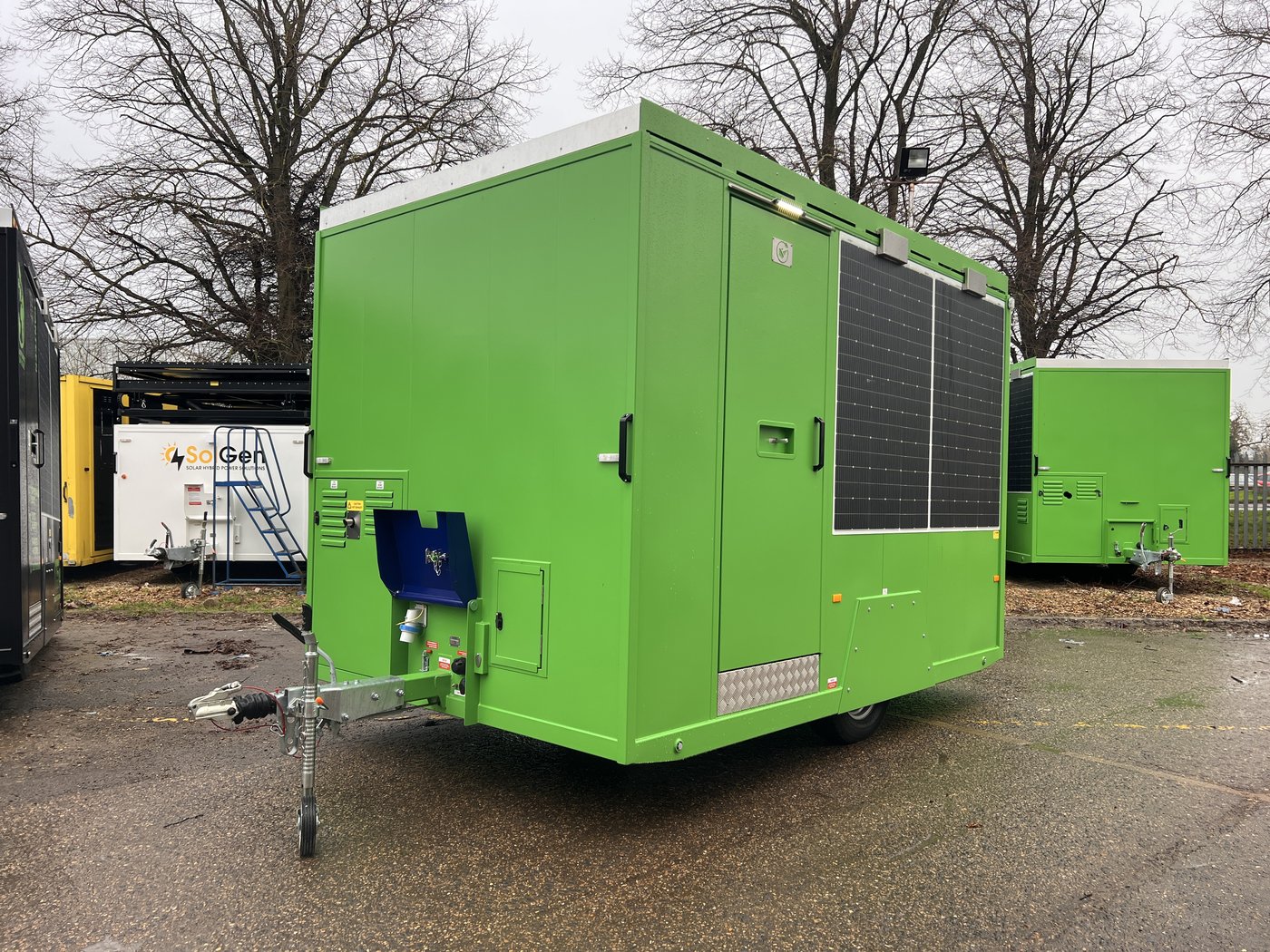 Fig. 2 — Front left view of the unit on road wheels, showing the tow hitch, jockey wheel, anti-vandal cover (closed), wall-mounted solar panels on the left side face, and blue fuel compartment