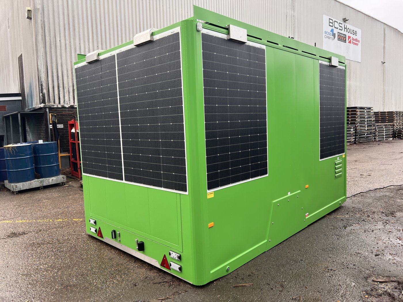 Fig. 43 — Full unit from rear left, photographed at the BCS factory prior to delivery. The unit is deployed on its hydraulic rams with all six wall-mounted solar panels clearly visible on the rear face. The access door panels and the generator compartment cover are visible on the left side. The anti-vandal cover flap is in the raised/open position