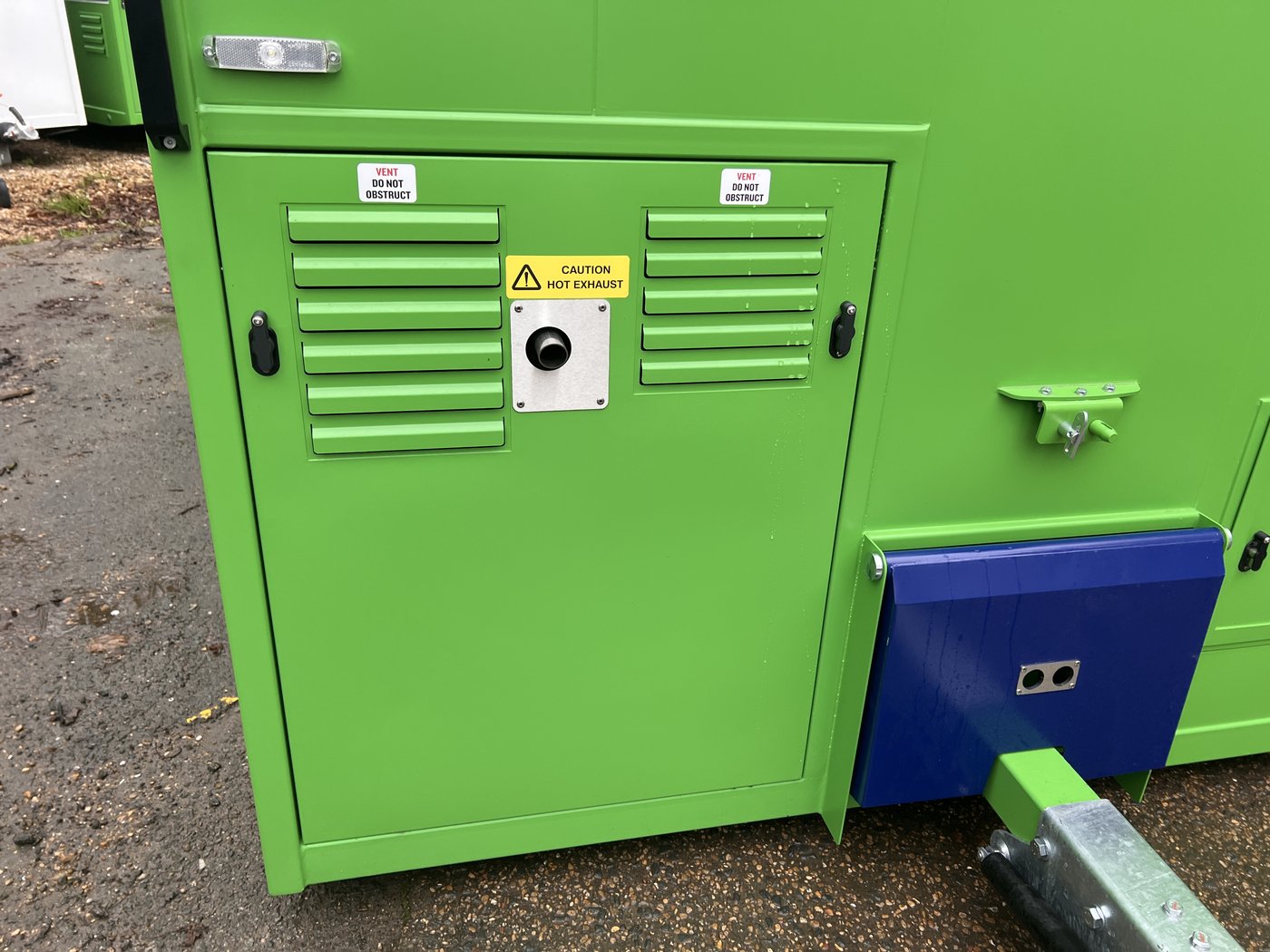 Fig. 23 — Generator compartment with the access cover fitted, showing the louvred ventilation panels ('VENT DO NOT OBSTRUCT'), the yellow 'CAUTION HOT EXHAUST' warning label, and the generator E-stop button in the centre of the cover. The blue fuel compartment is visible to the right