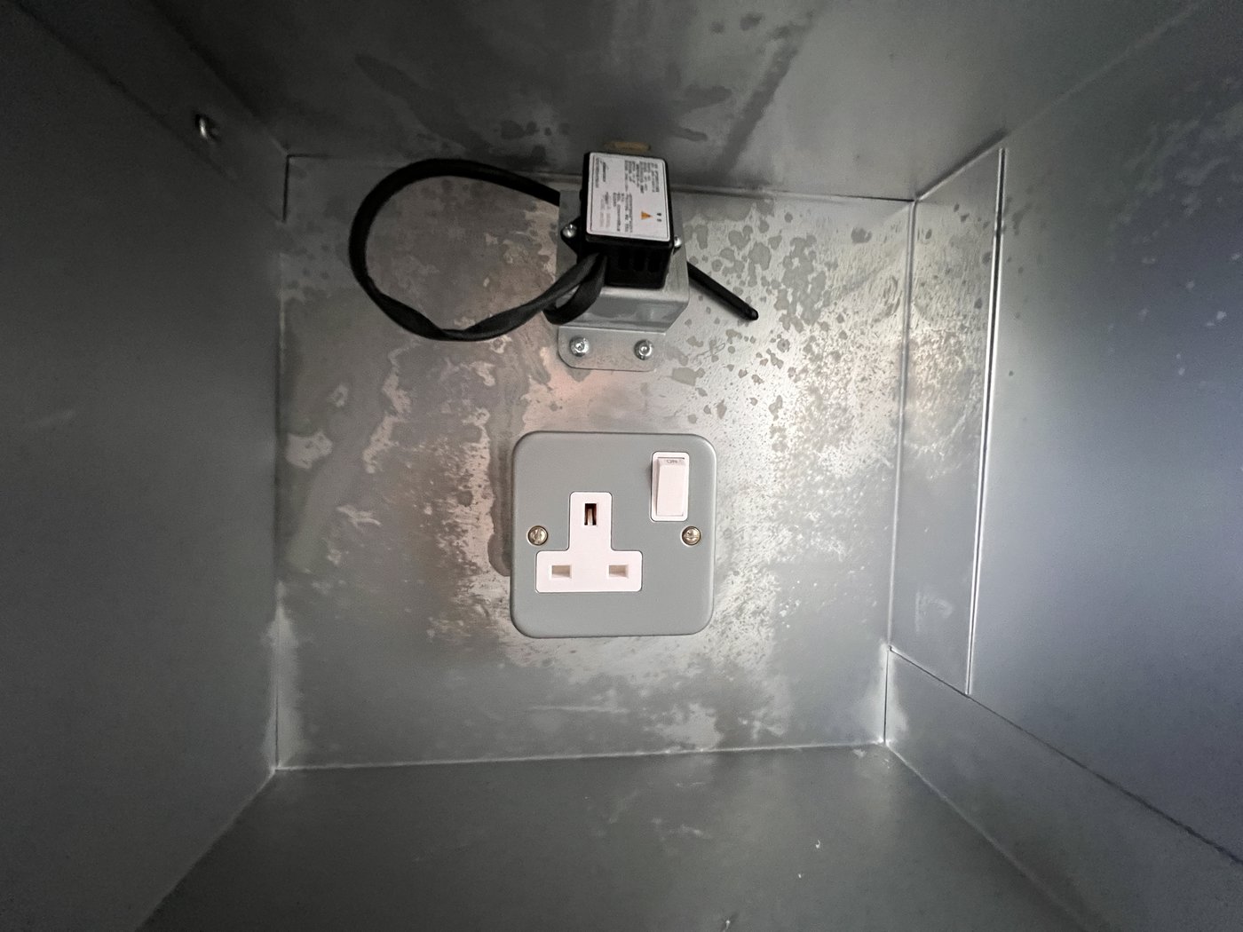 Fig. 19 — Interior of a single charging compartment showing the 230V switched socket on the rear wall and the fire suppression module (black box with warning label) mounted above it. The galvanised steel interior minimises combustible material