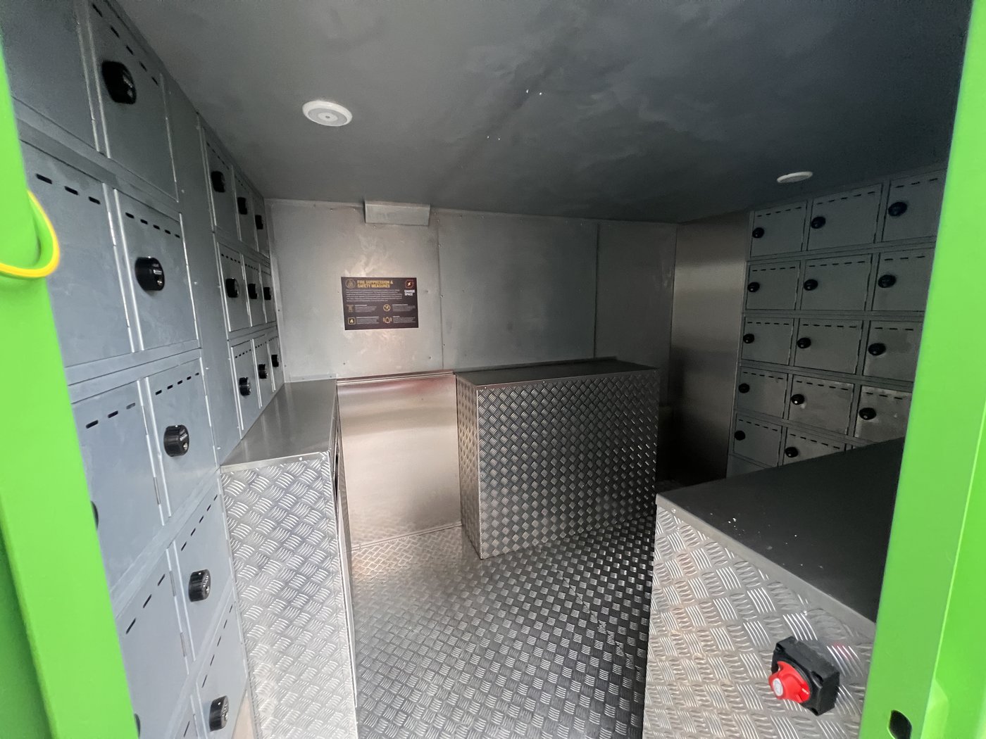 Fig. 13 — View from the access doorway showing charging compartments on both walls, the central counter unit, fire safety information panel on the far right wall, and the red E-stop mushroom button visible at the lower right of the door frame