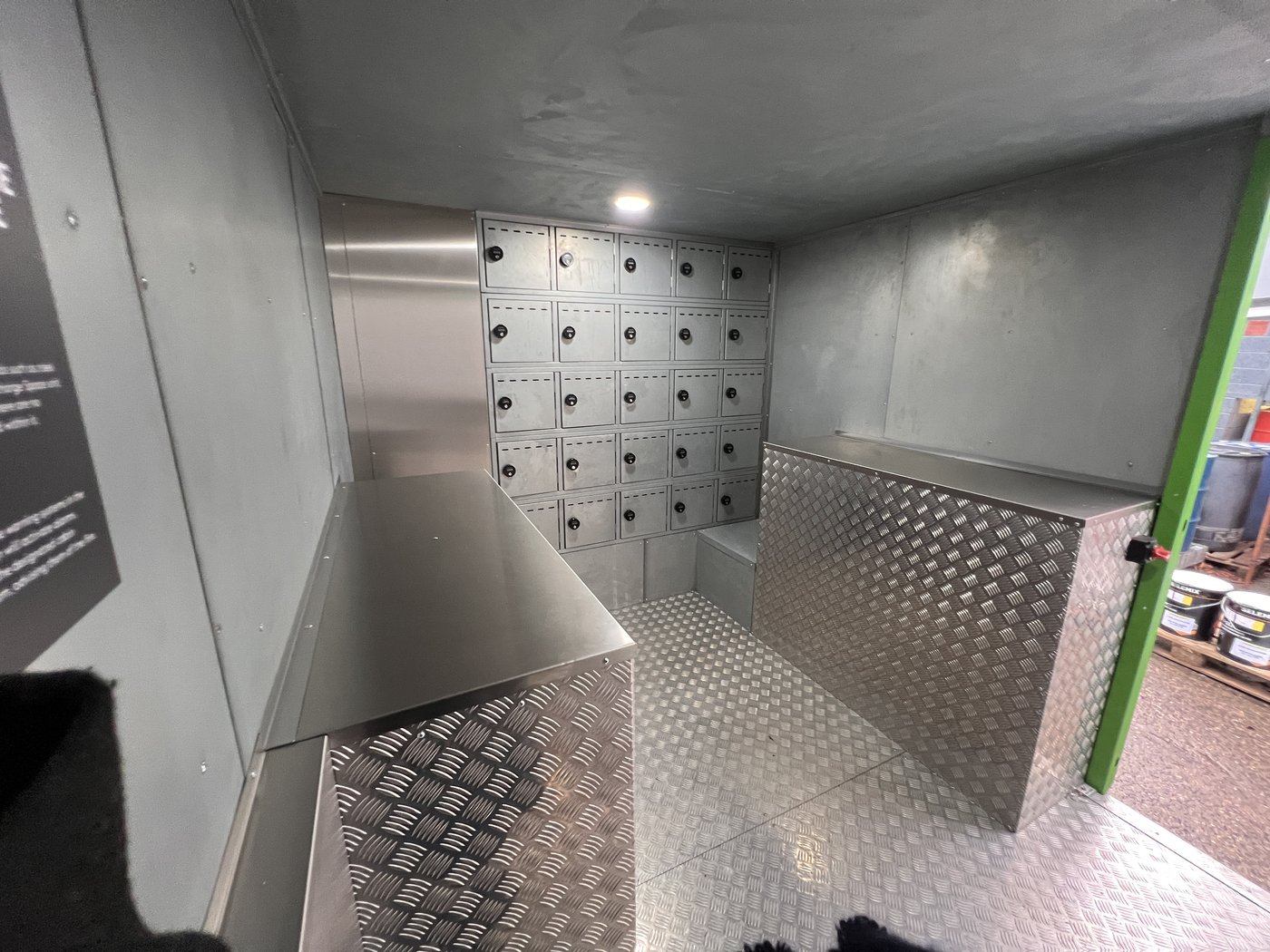 Fig. 10 — Interior overview from the front right corner, showing the stainless steel locker bank (approximately 25 compartments visible), chequerplate aluminium floor, and stainless steel shelf unit on the left side. The access door is open to the right