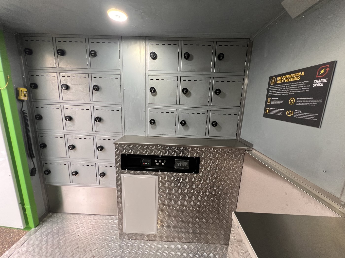Fig. 12 — Rear interior wall showing the left-hand bank of charging compartments with KitLock combination locks, the central counter unit housing the display screen, and the Fire Suppression and Safety Measures information panel mounted on the right wall