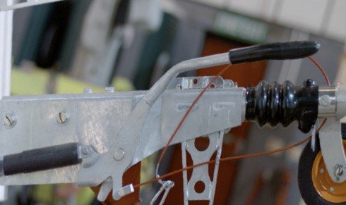 Fig. 64 — Overrun coupling close-up showing the brake cable routing (orange cable), handbrake lever pivot, and the compensator bar connecting both brake cables. The red cable annotation shows correct alignment