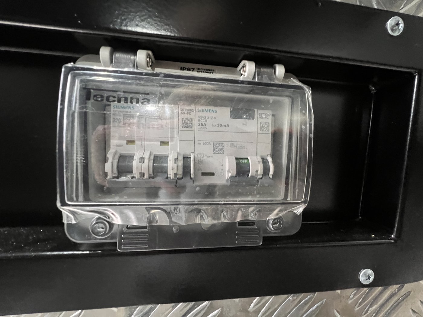 Fig. 25 — The RCD distribution board, housed in an IP67-rated enclosure with a clear hinged cover. The board contains a Siemens RCBO (left) and MCG breaker (right), with all conductors entering from below through a sealed gland plate
