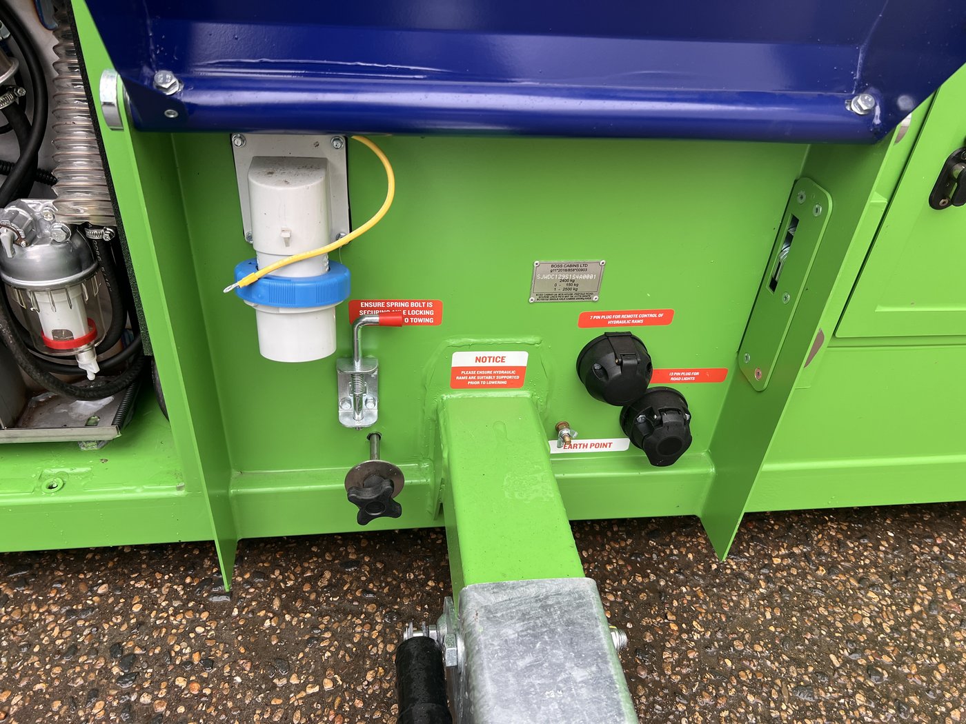 Fig. 22 — Under the anti-vandal cover: the white external AC inlet socket (CEE type, with blue sealing cap), fuel filter/water separator (left), and the orange E-stop mushroom button. Tow hitch hardware is visible below